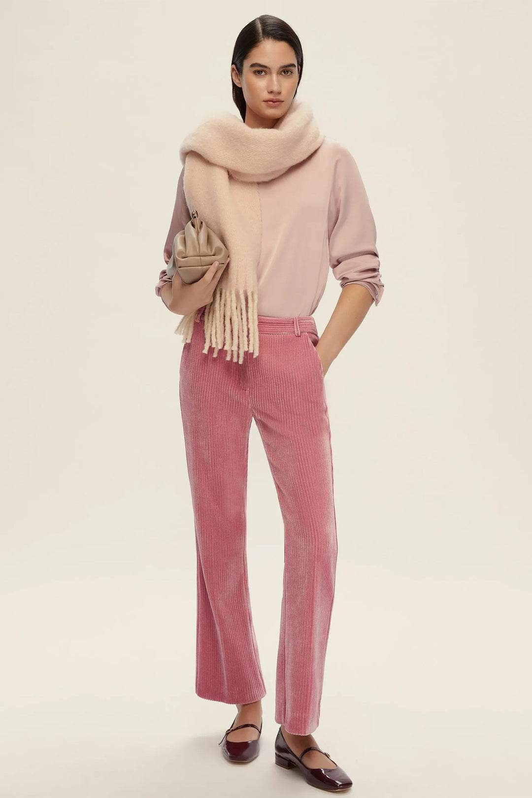 Woman wearing a pink sweater, scarf, and pants with a beige handbag on a white background