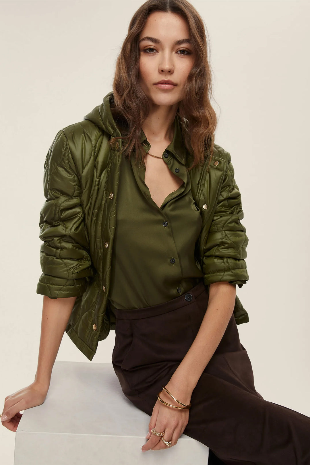 Woman wearing an olive green jacket over a shirt, sitting on a white surface.