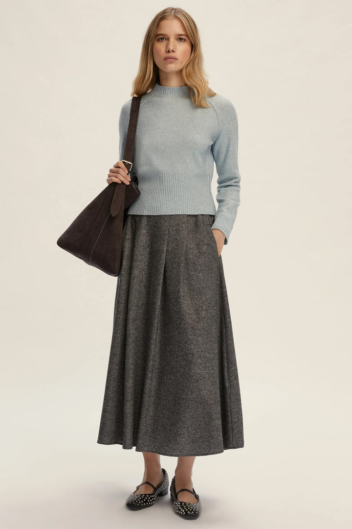 Woman wearing a light blue sweater and gray skirt holding a brown bag on a beige background