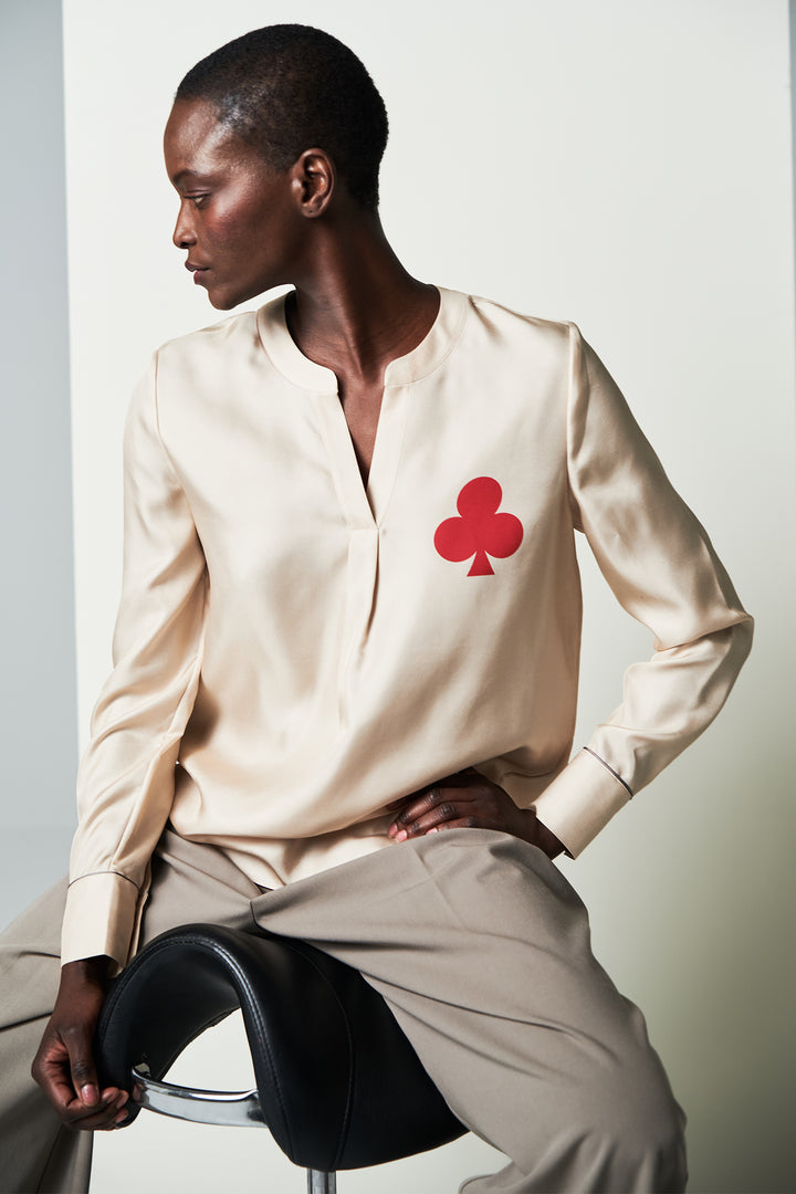 Person wearing a cream shirt with a red clover symbol, sitting on a black stool against a white background