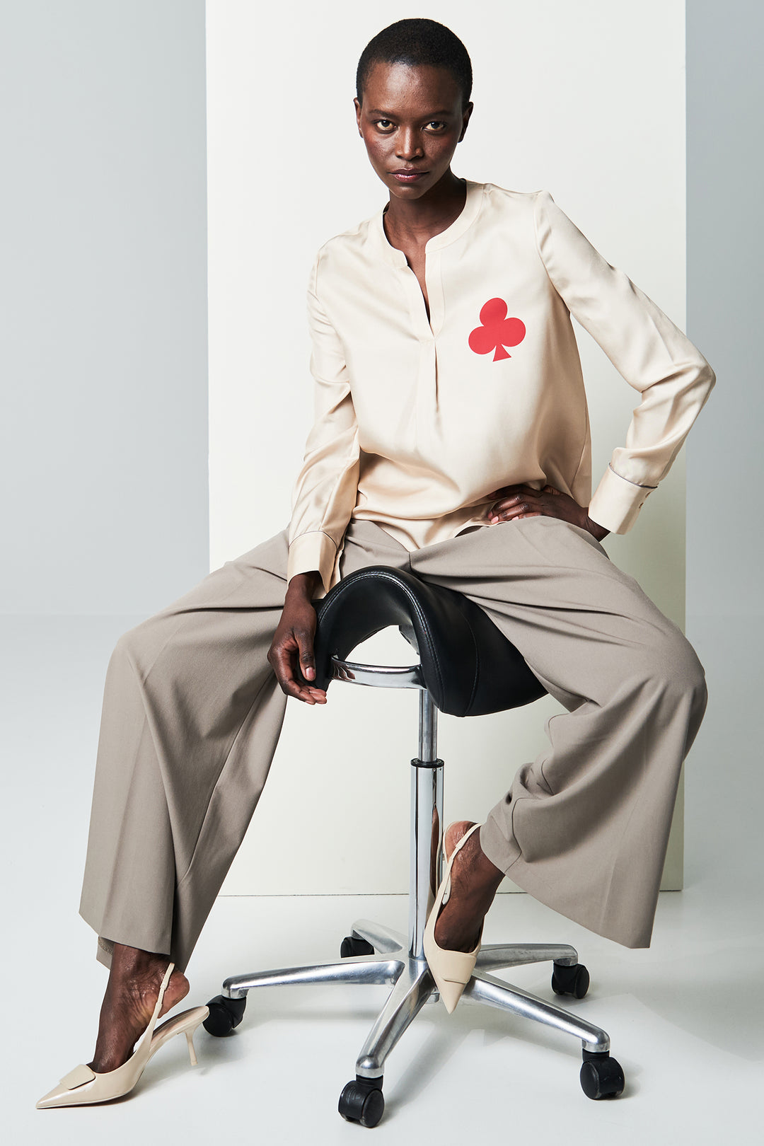 Person sitting on a stool wearing a beige outfit with a red clover symbol.