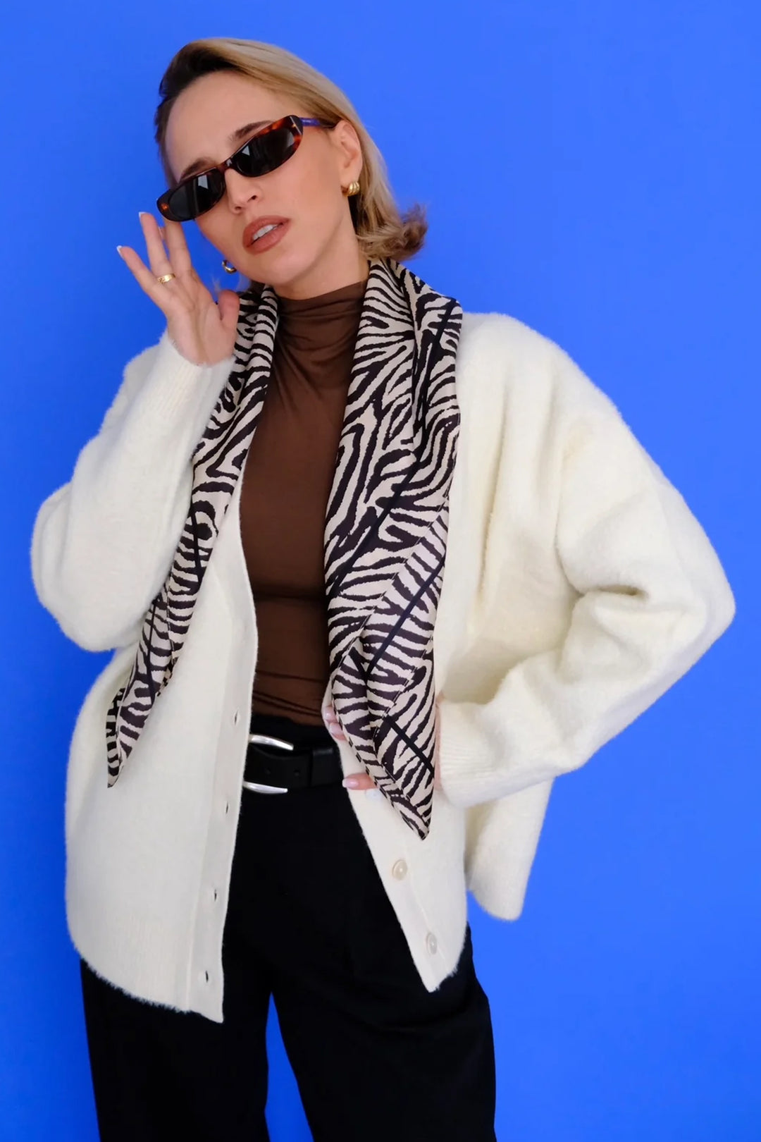 Person wearing a white coat, brown top, black pants, and a zebra print scarf against a blue background
