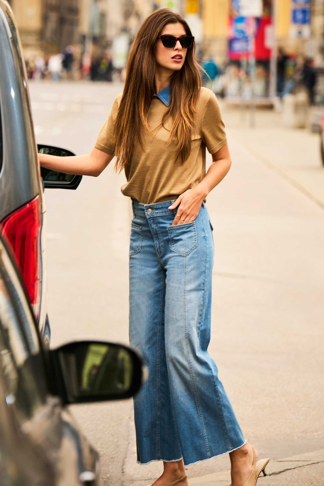 Woman in a beige top and blue jeans getting into a car on a city street.