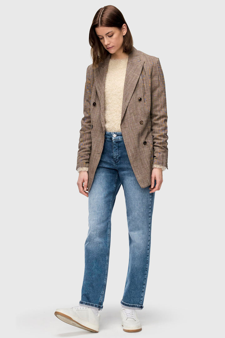 Woman wearing a brown blazer, beige sweater, and blue jeans on a white background