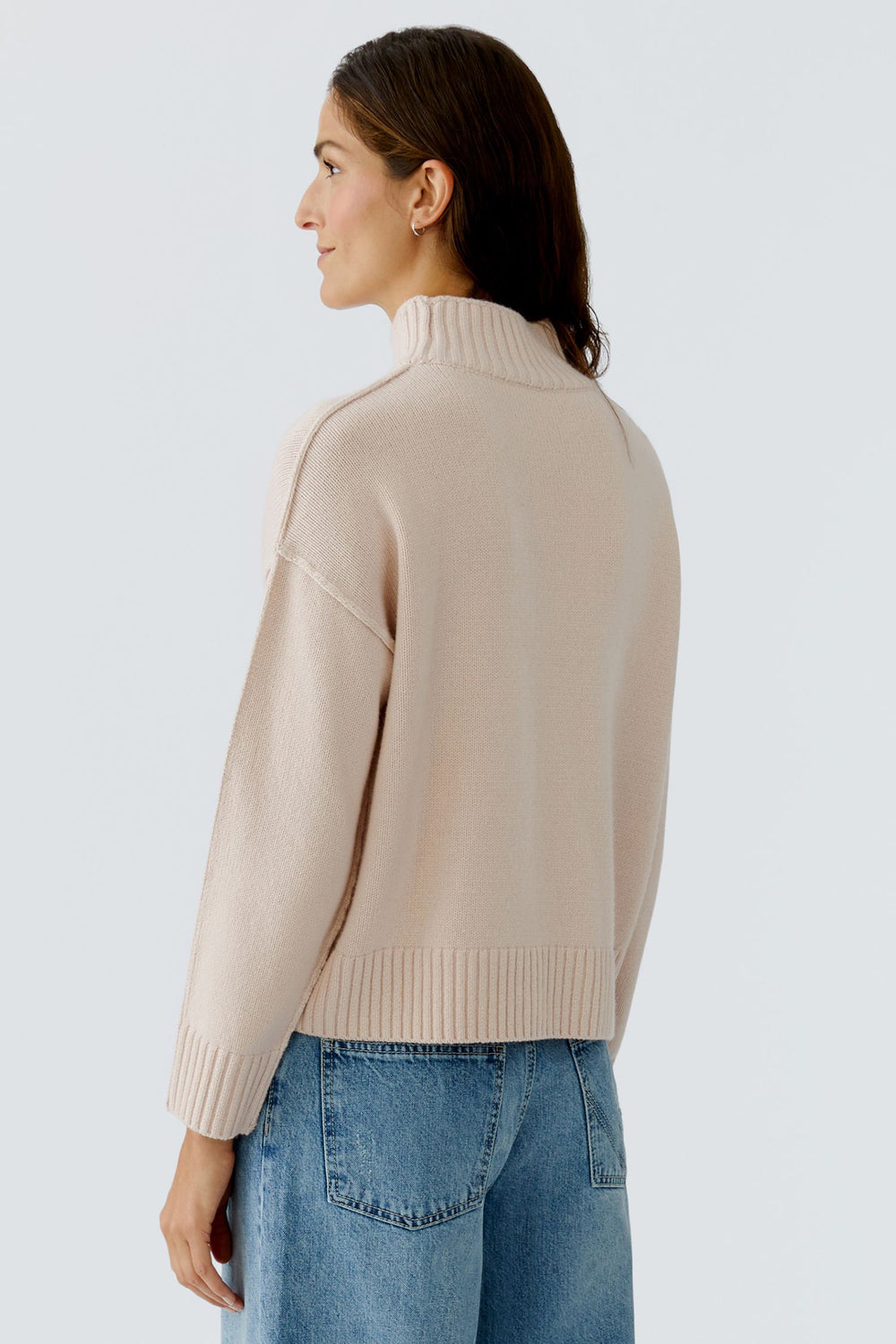 Woman wearing a beige turtleneck sweater and blue jeans on a light gray background