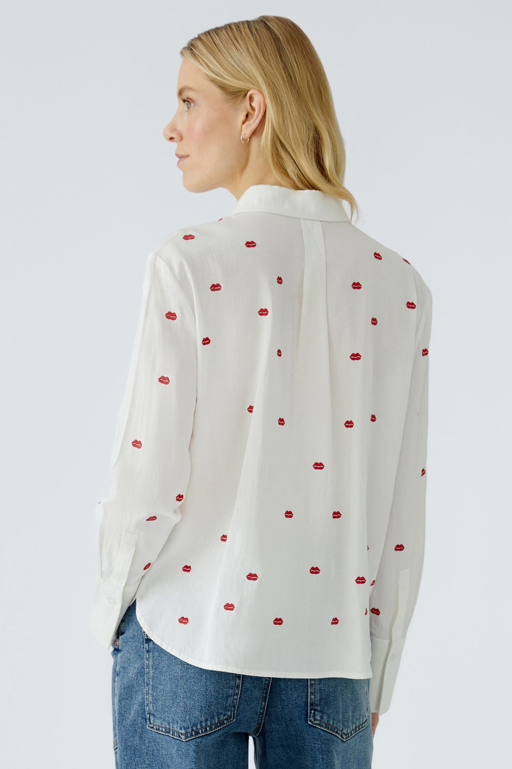 Woman wearing a white blouse with red patterns and blue jeans on a light gray background