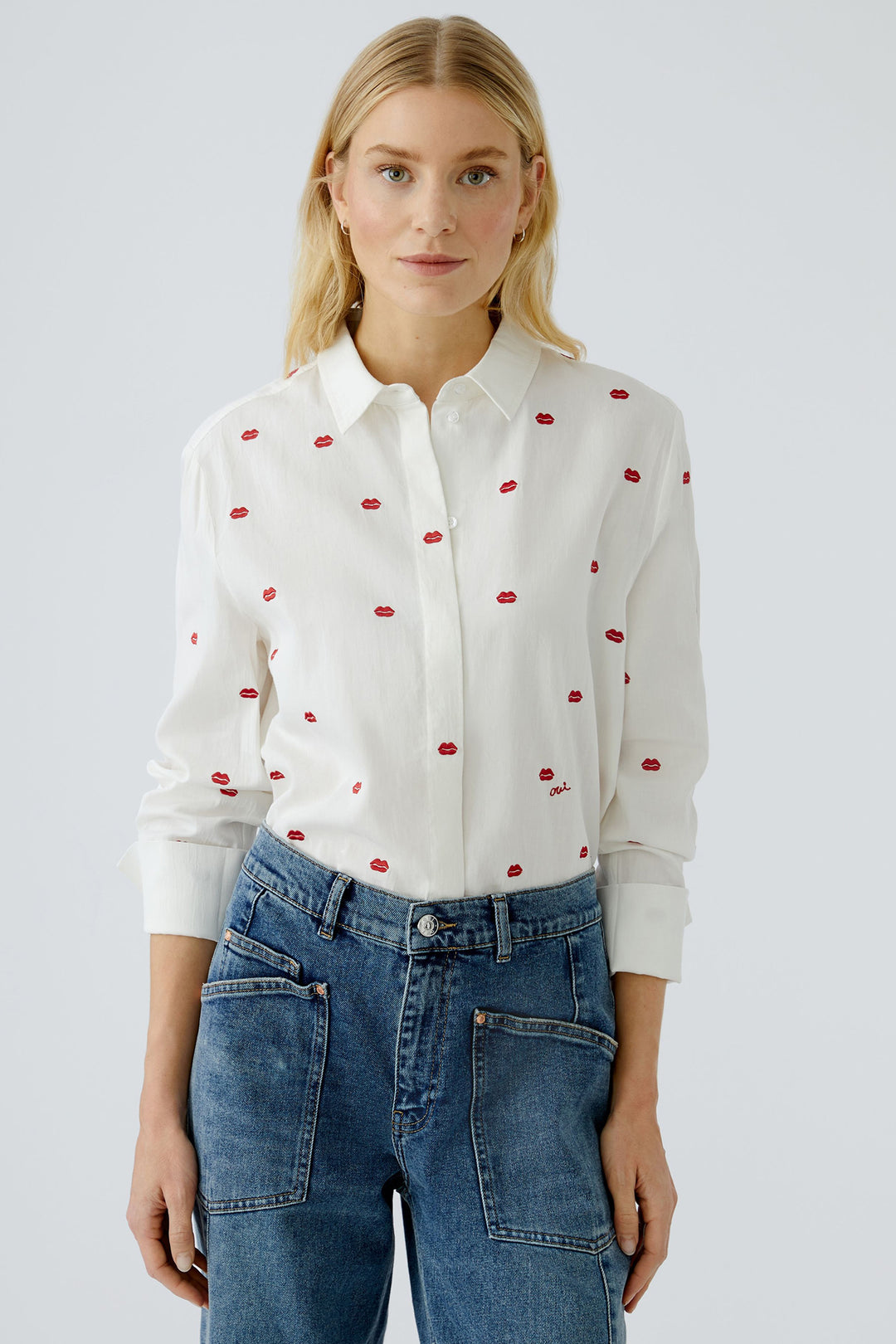 Woman wearing a white blouse with red patterns and blue jeans on a light gray background
