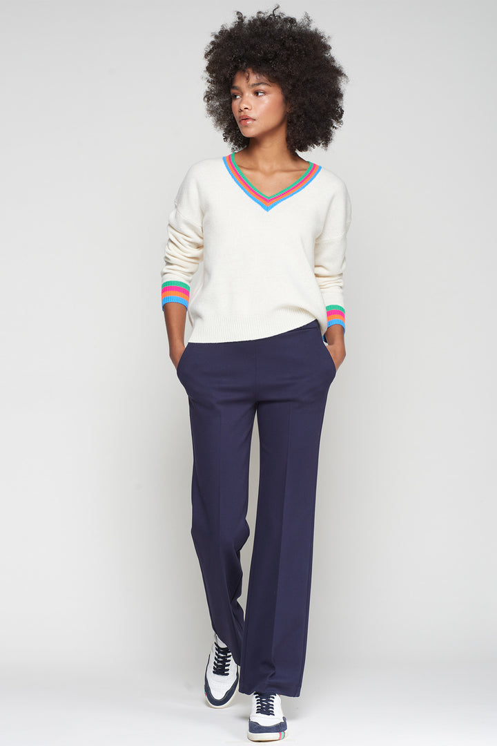 Woman wearing a white sweater with colorful trim and navy pants on a light gray background