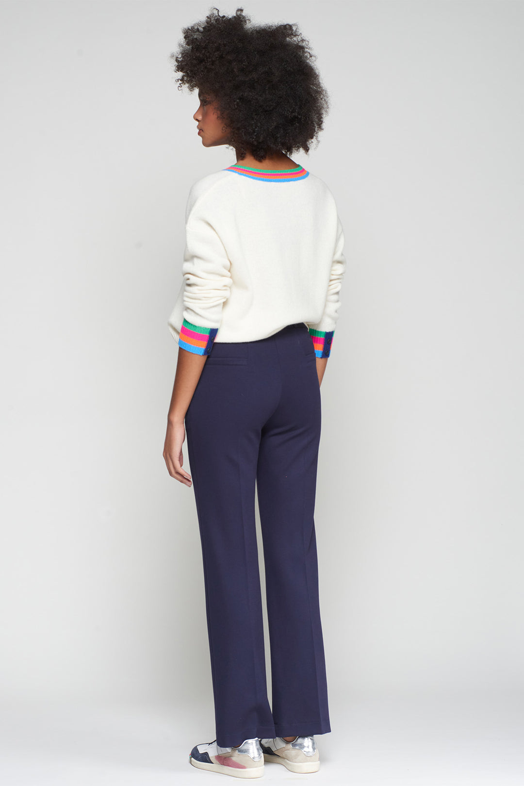 Person wearing a white sweater with colorful stripes and navy pants on a plain background