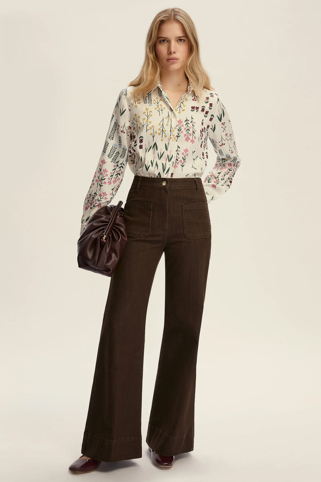Woman wearing a floral blouse and brown pants with a brown handbag on a beige background