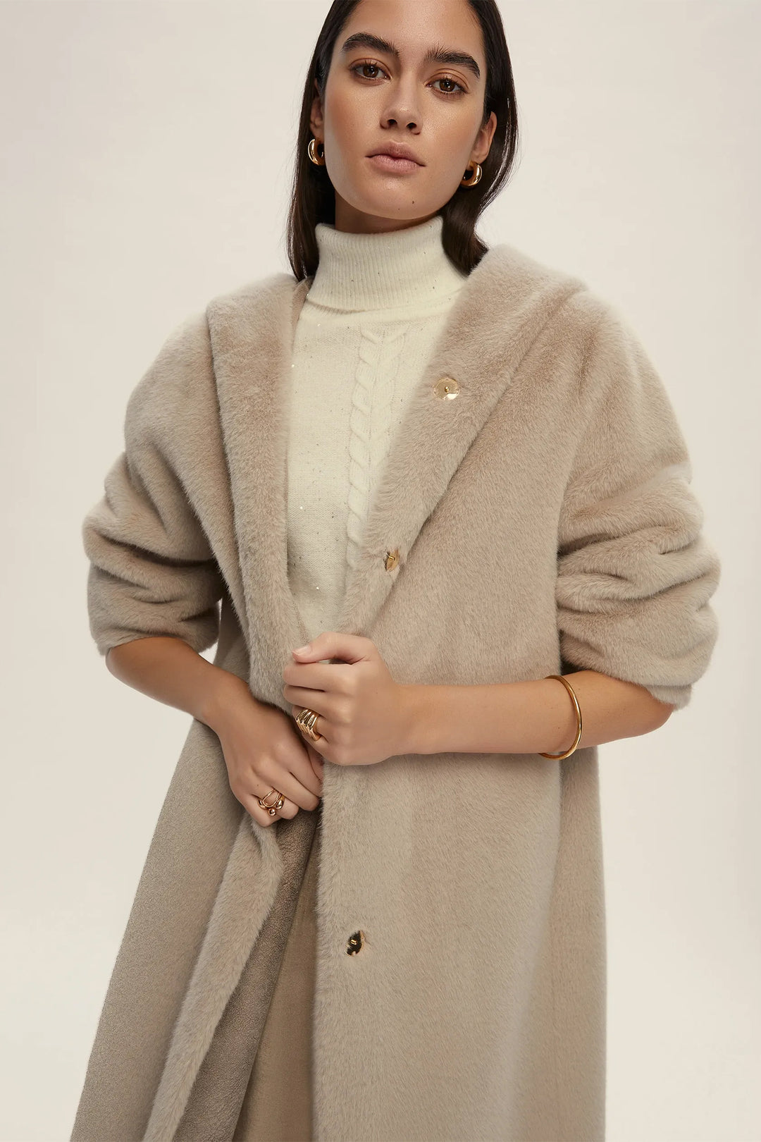 Woman wearing a beige coat over a white turtleneck sweater on a plain background
