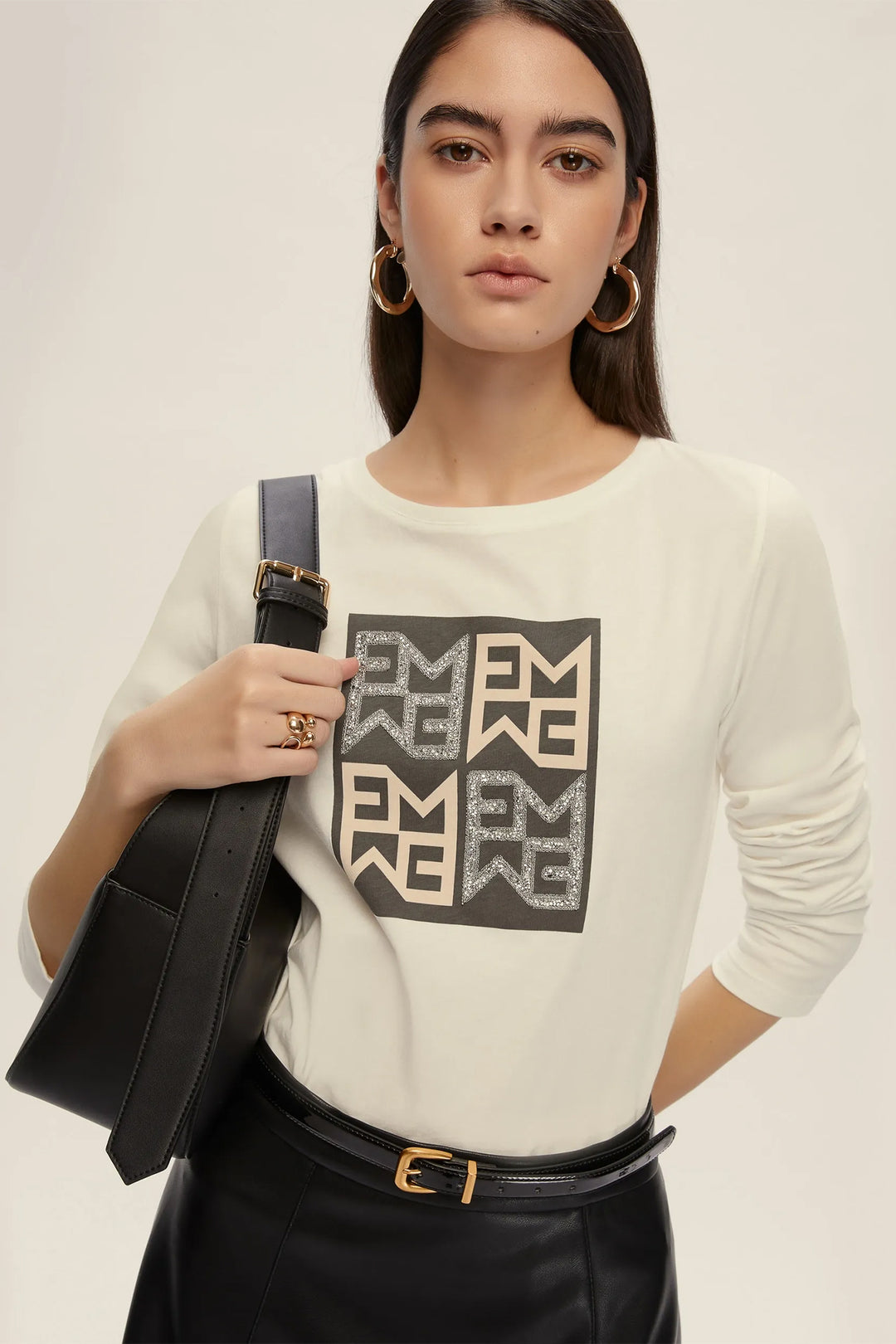 Woman wearing a white sweater with a graphic design, holding a black bag on a plain background