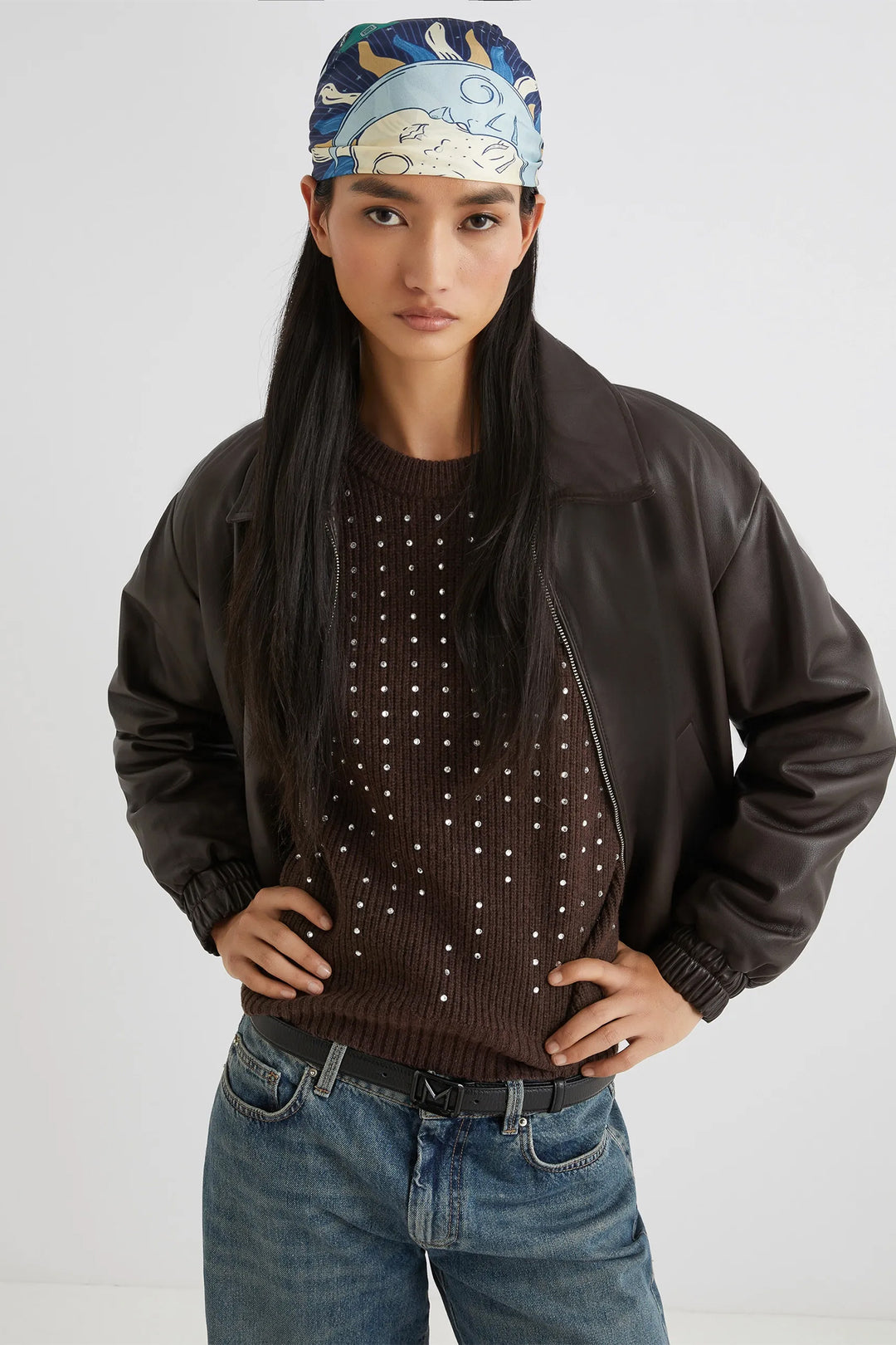 Person wearing a brown jacket, studded sweater, and blue jeans with a patterned headband on a white background