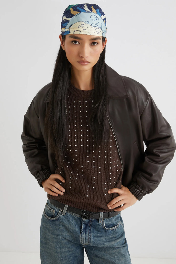 Person wearing a brown jacket, studded sweater, and blue jeans with a patterned headband on a white background