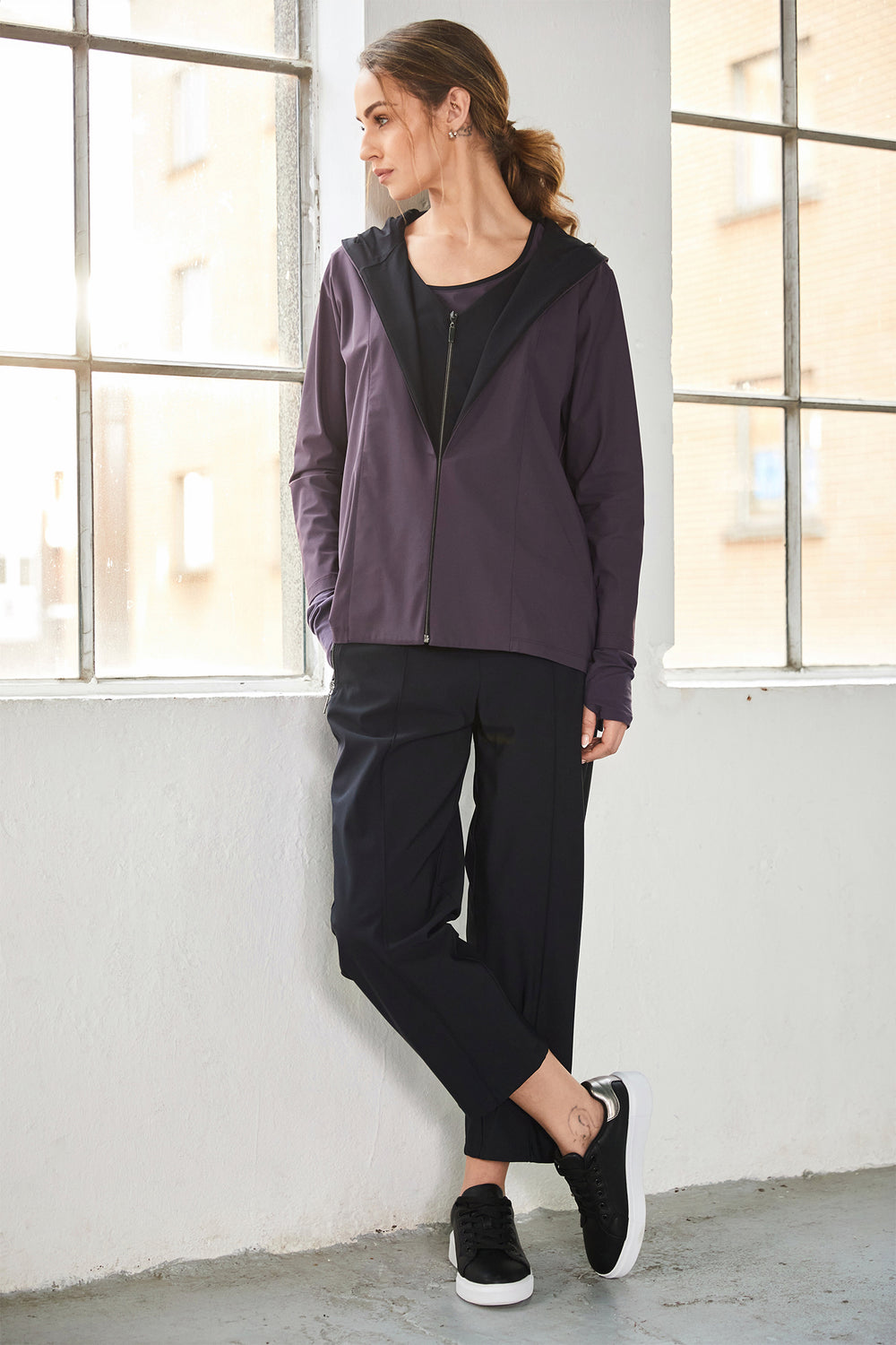 Woman wearing a dark purple jacket and black pants standing in a room with large windows.