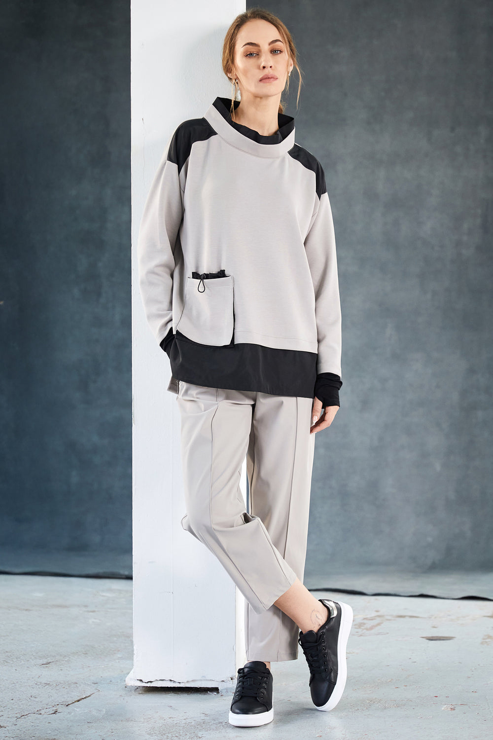 Person wearing a taupe and black outfit with a hood against a gray background