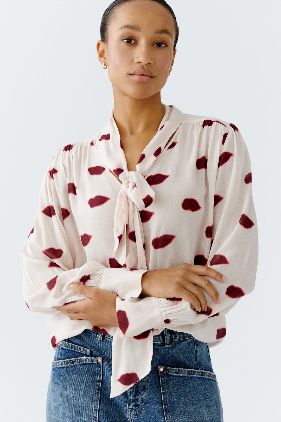 Woman wearing a white blouse with red heart patterns and blue jeans on a light gray background