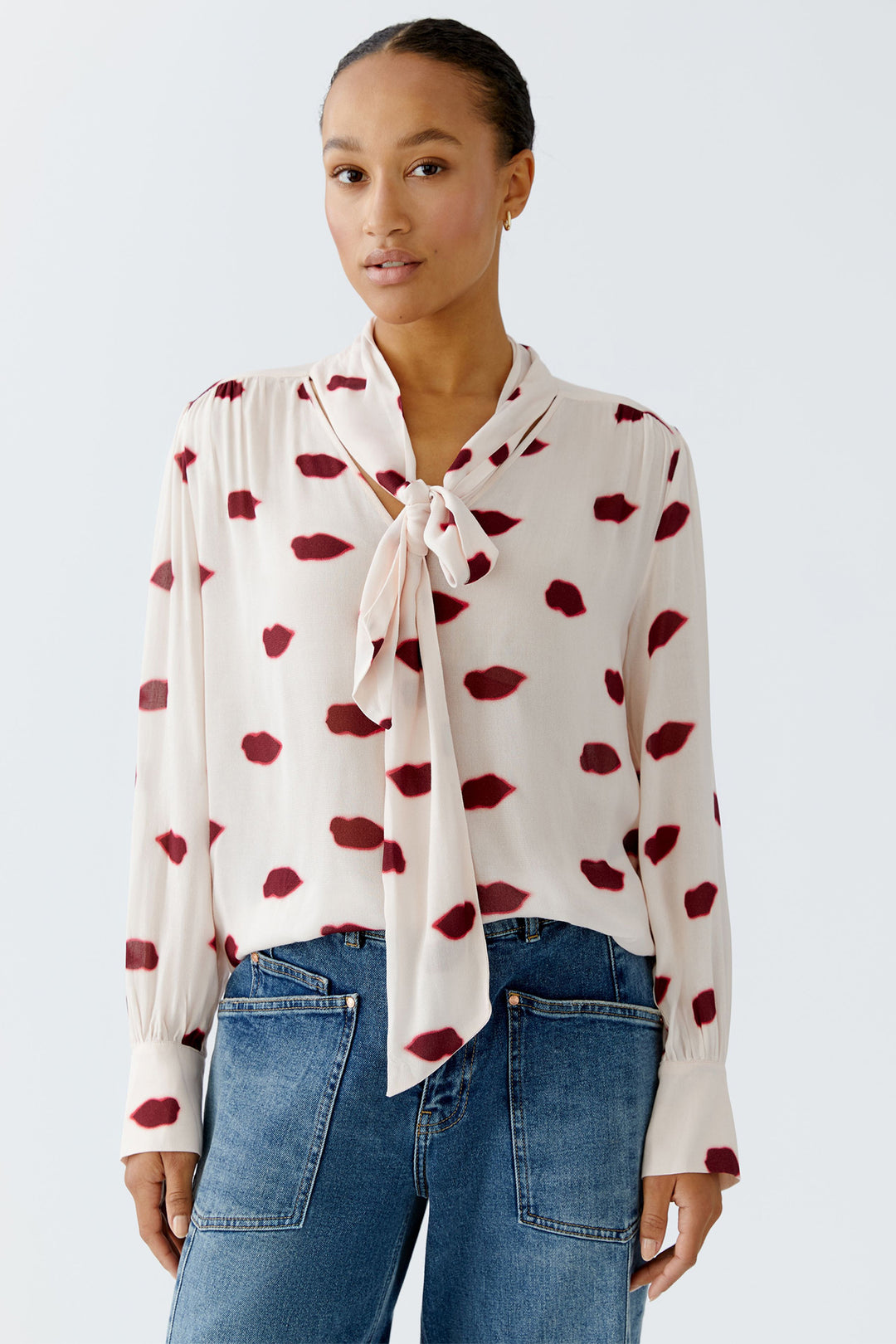 Woman wearing a white blouse with red pattern and blue jeans on a light gray background