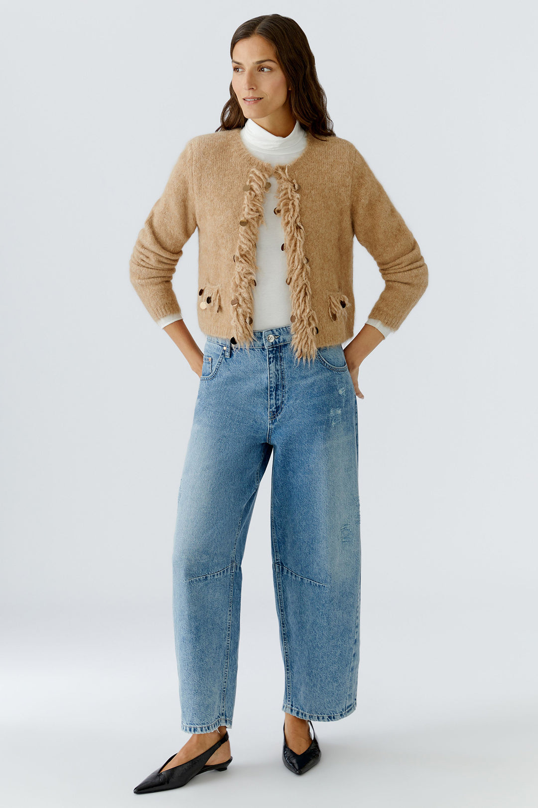 Woman wearing a beige cardigan with ruffled details, white shirt, and blue jeans on a light gray background