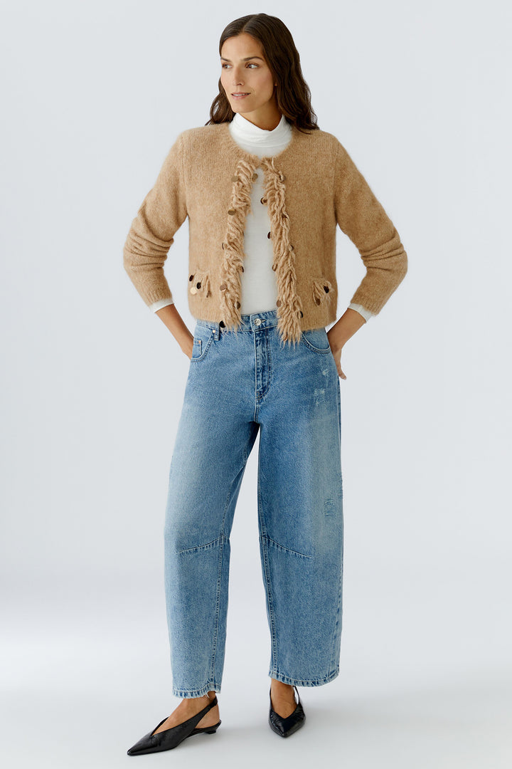 Woman wearing a beige cardigan with ruffled details, white shirt, and blue jeans on a light gray background