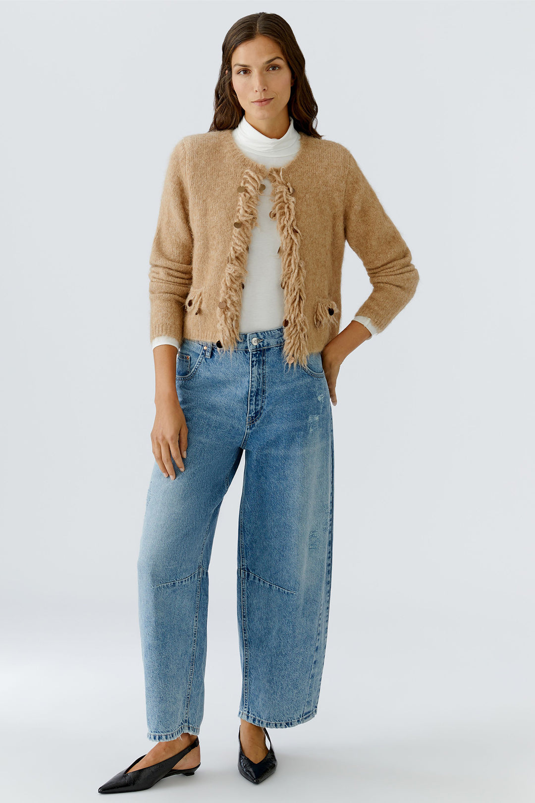 Woman wearing a beige cardigan with fringe, white sweater, and blue jeans on a light gray background